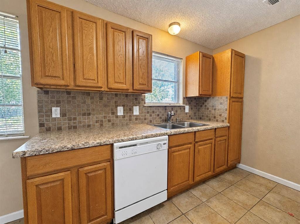 2418 Fulton Drive, Mesquite, TX 75150 Kitchen featuring wood cabinetry, stone-patterned laminate countertops, a mosaic tile backsplash, a double-basin stainless steel sink, and ceramic tile flooring