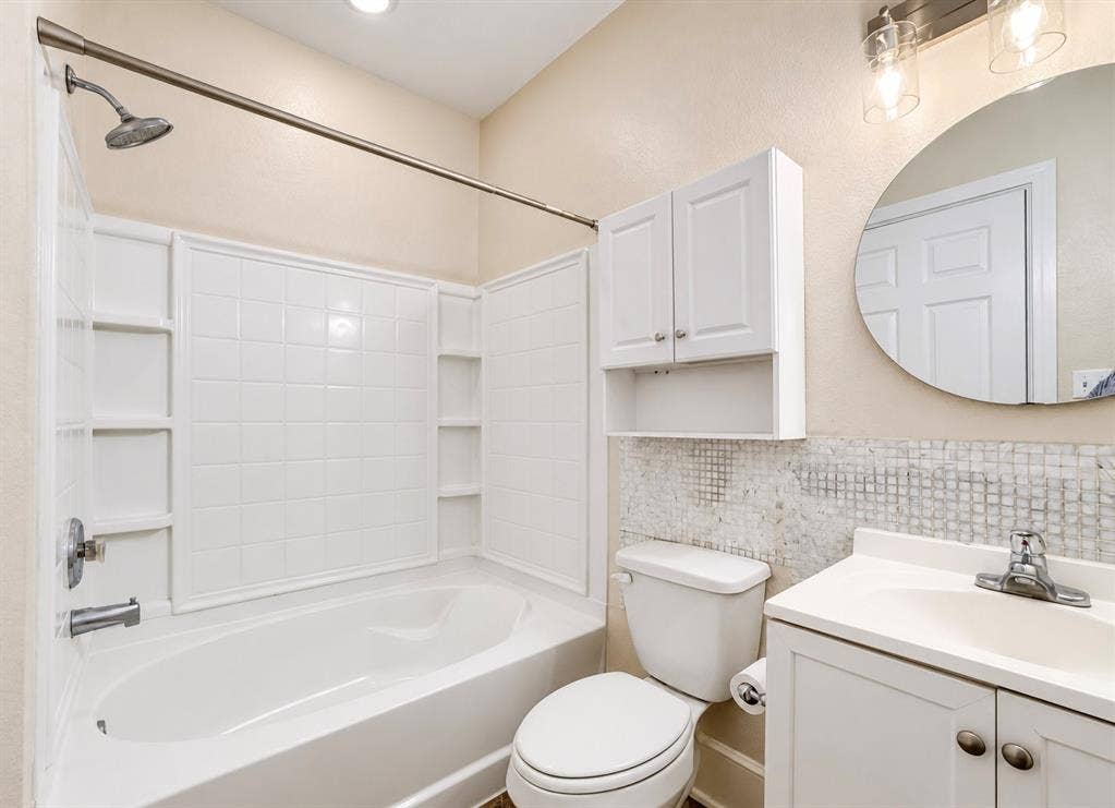 4227 Delano Place, Dallas, TX 75204 Bathroom featuring a white tub-and-shower combination with integrated shelving, white vanity cabinet with a solid-surface top, circular wall-mounted mirror, and wall-mounted storage cabinetry