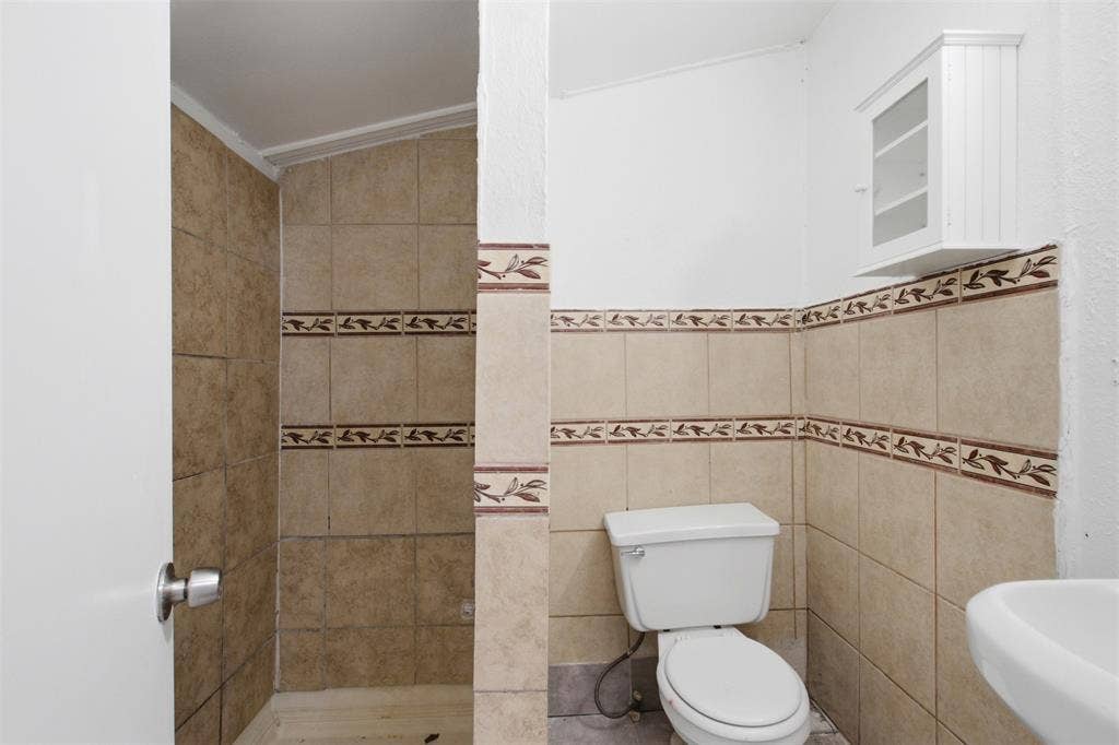 2009 S 1st Street, Garland, TX 75041 Full-height ceramic wall tiling featuring a decorative border, wall-mounted storage cabinet, and white porcelain toilet