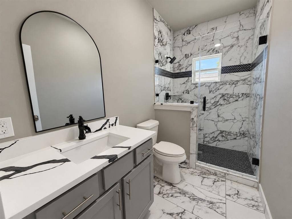 707 N Blanche Street, Terrell, TX 75160 Bathroom featuring a gray vanity with a stone-finish countertop, arched-top mirror, walk-in shower with large-format tile surrounds, and matte black fixtures