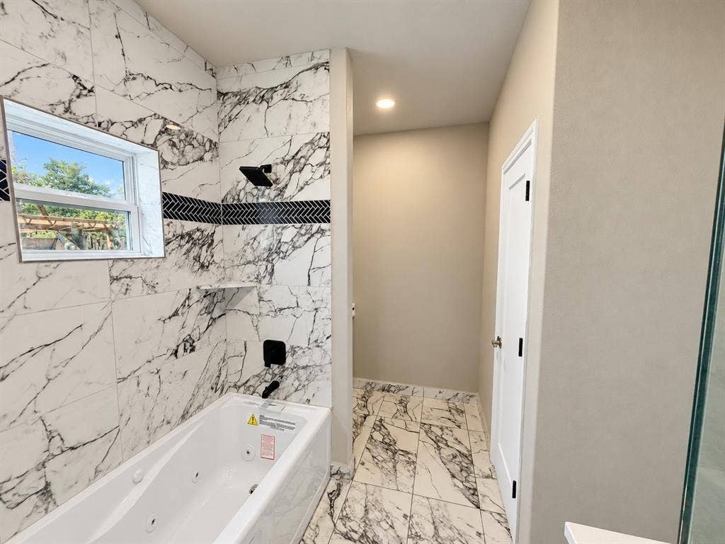 707 N Blanche Street, Terrell, TX 75160 Jetted soaking tub with large-format veined tile surround, matte black rainfall shower fixture, decorative herringbone tile inlay, and matching stone-patterned tile flooring