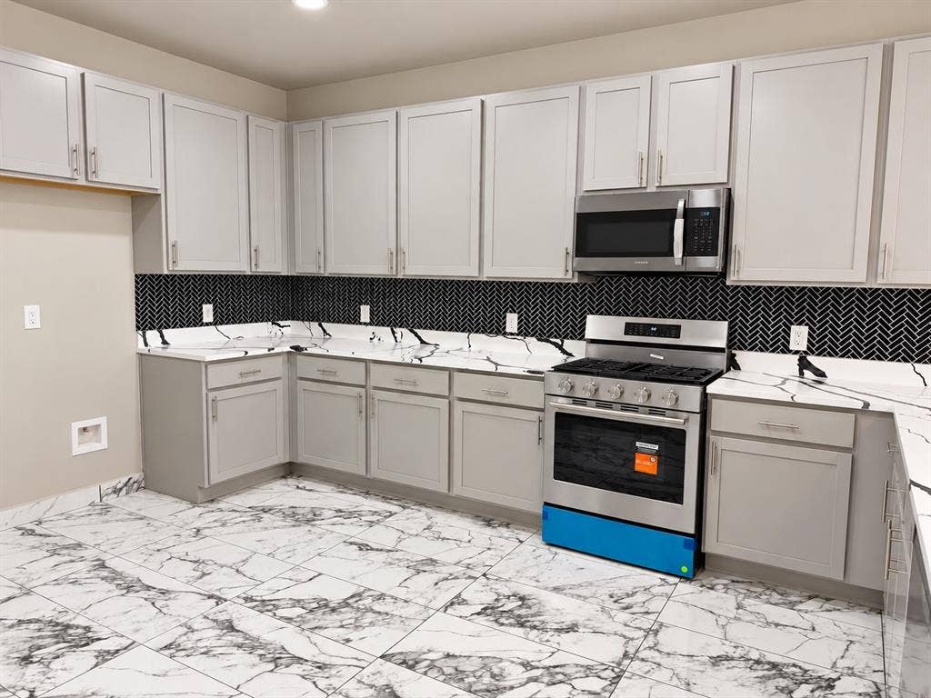 707 N Blanche Street, Terrell, TX 75160 Kitchen featuring light gray shaker-style cabinetry, patterned stone-finish countertops, black herringbone tile backsplash, stainless steel appliances, and large-format veined tile flooring