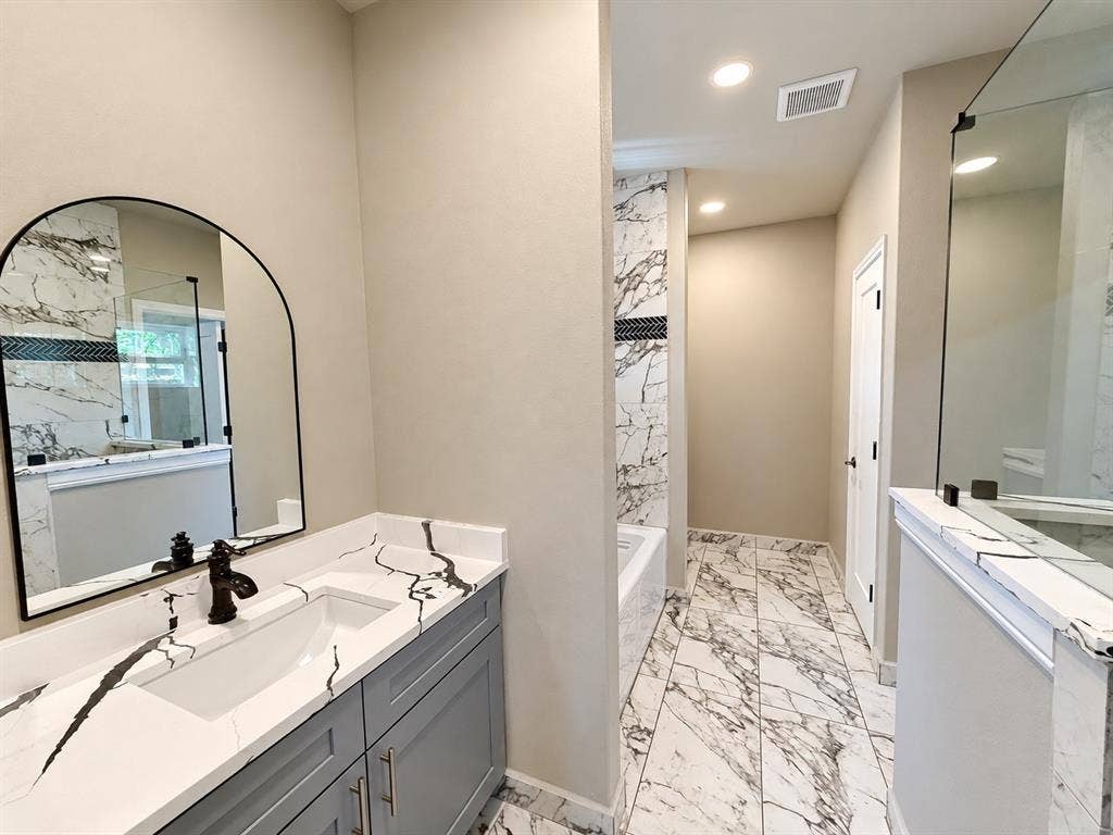 707 N Blanche Street, Terrell, TX 75160 Bathroom featuring gray shaker-style cabinetry, white countertops with dark veining, an arched wall mirror, large-format tile flooring, and a walk-in shower with glass enclosure