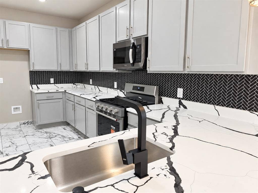 707 N Blanche Street, Terrell, TX 75160 Kitchen featuring gray shaker-style cabinetry, high-contrast herringbone tile backsplash, white stone-patterned countertops, stainless steel appliances, and a matte black gooseneck faucet