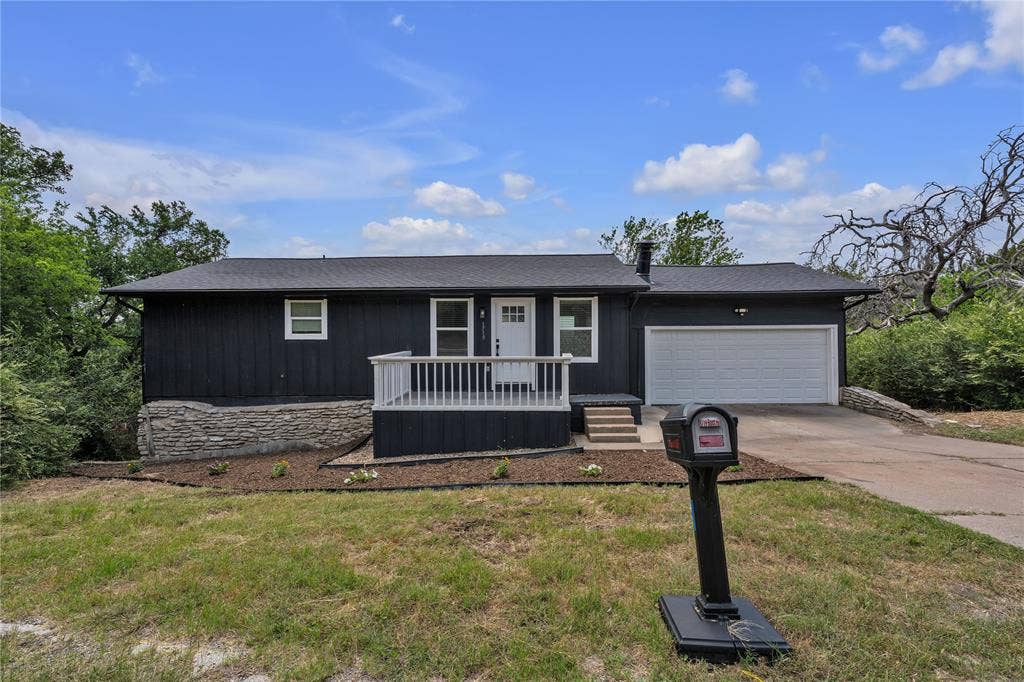 1719 Boot Hill Road, Granbury, TX 76049 Single-story exterior featuring dark vertical siding, an attached garage, a raised deck with white railing, and a stone veneer foundation