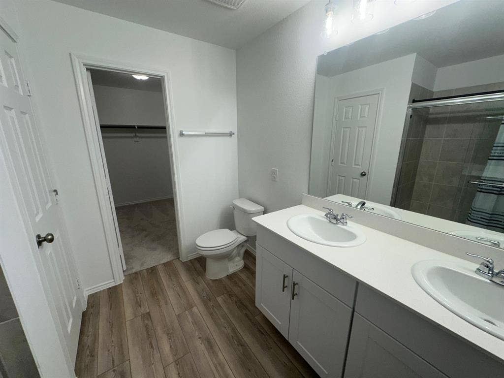 821 POPPY COVE Lane, Princeton, TX 75407 Bathroom featuring a dual-sink vanity with a solid-surface countertop, white cabinetry with brushed metal hardware, wood-finish flooring, and a full-width mirror