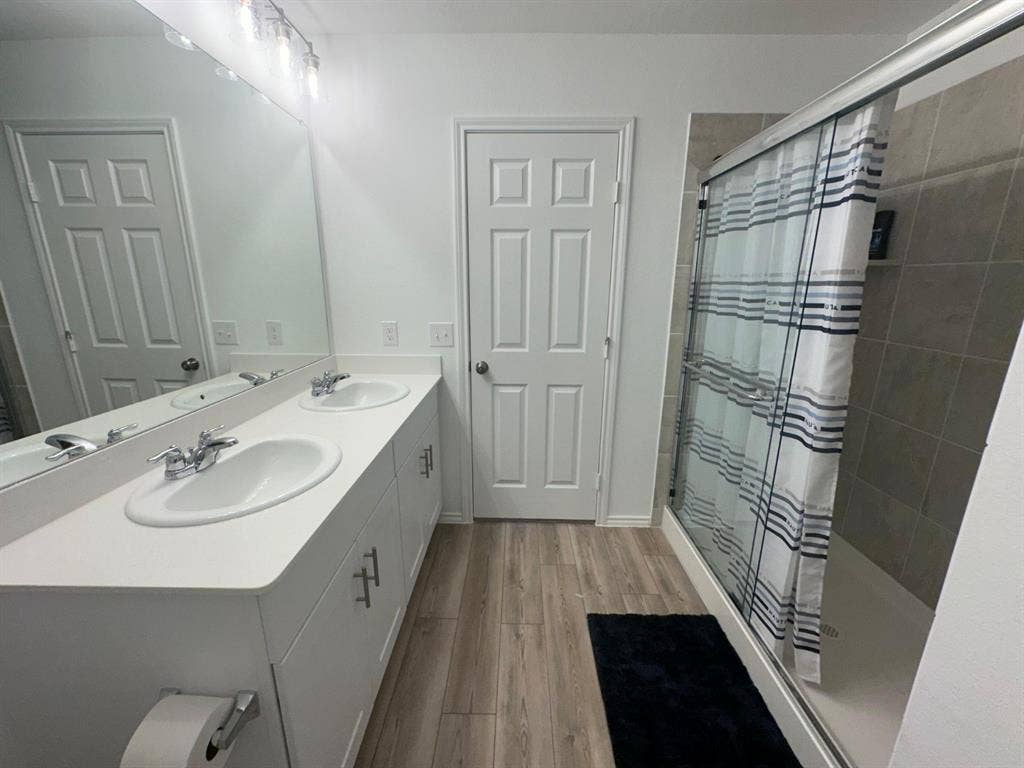 821 POPPY COVE Lane, Princeton, TX 75407 Dual-sink vanity with white countertop, full-width vanity mirror, wood-finish flooring, and glass-enclosed shower featuring tiled surround