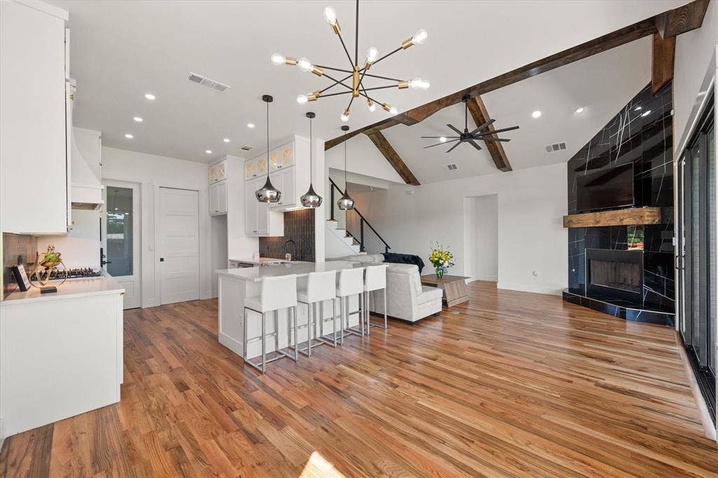 122 Yvonne Street, Red Oak, TX 75154 Open-concept interior featuring vaulted ceilings with decorative wood beams,Oak wood flooring, a kitchen island with pendant lighting, and a floor-to-ceiling fireplace with a black tile surround and a wood mantel