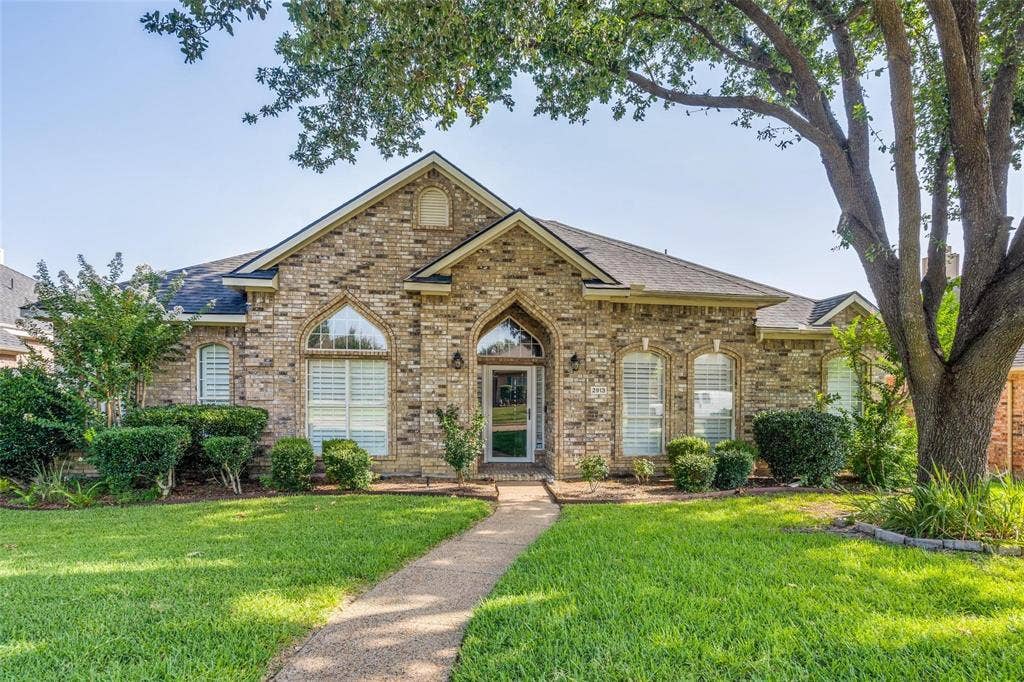 2913 Bellerive Drive, Plano, TX 75025 Brick exterior featuring arched window detailing, a steep-pitched roofline, and a concrete pathway leading to the front entrance, set amidst a landscaped front yard with mature trees