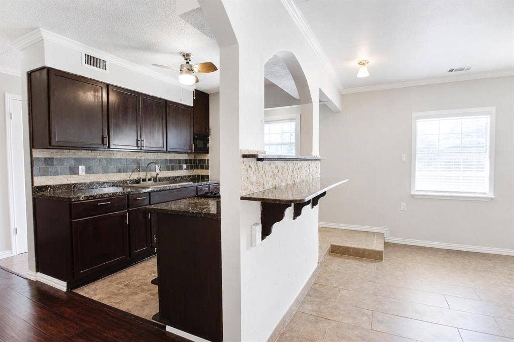 2708 N Avenue, Plano, TX 75074 Kitchen featuring dark cabinetry, stone-surface countertops, stone tile backsplash, breakfast bar with stone veneer detailing, and arched wall opening