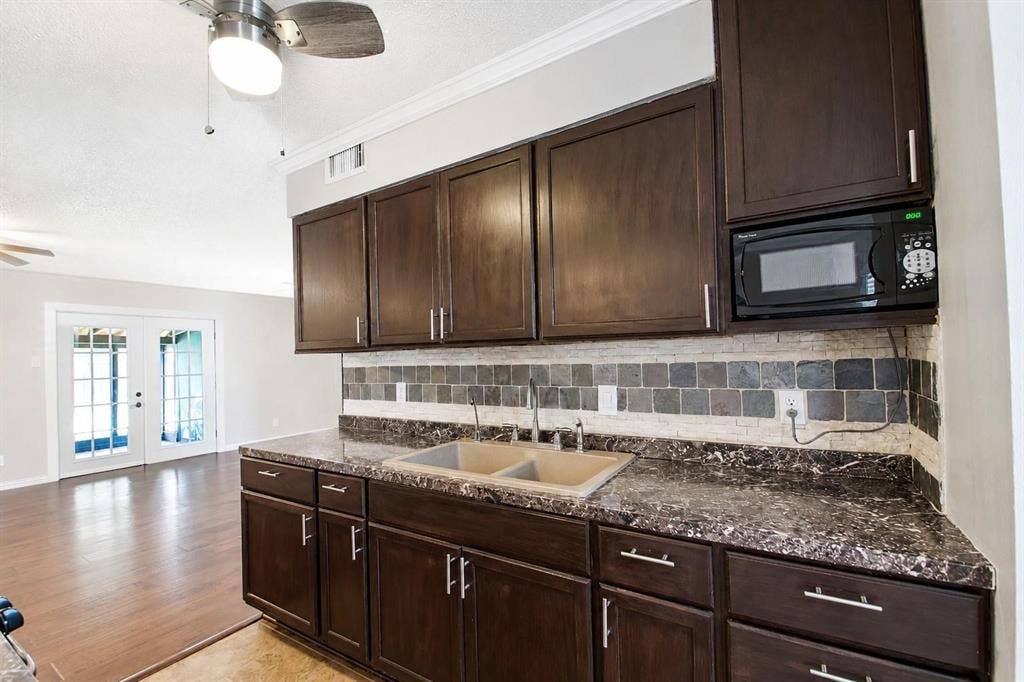 2708 N Avenue, Plano, TX 75074 Kitchen featuring dark-finish cabinetry, stone-patterned countertops, a dual-basin sink, multi-tone tile backsplash, and a built-in microwave