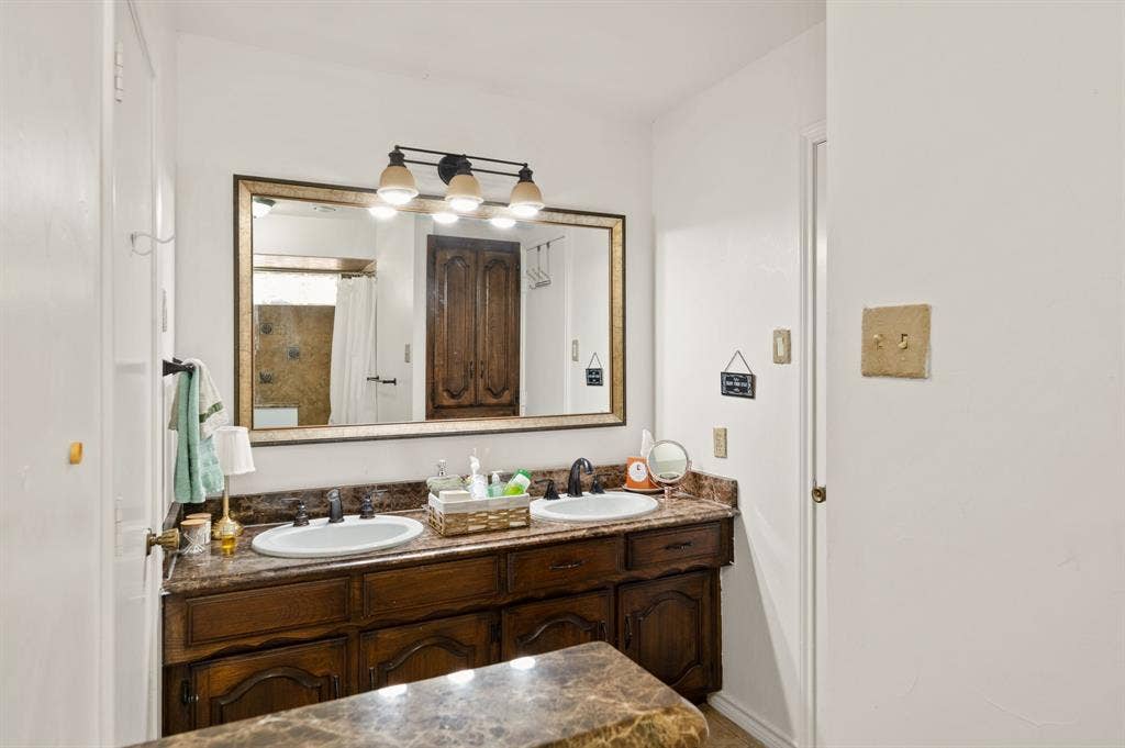 621 Harvest Glen Drive, Richardson, TX 75081 Dual-basin vanity featuring stone-finish countertops, dark wood cabinetry, integrated oval sinks, and a large framed wall mirror with a multi-bulb light fixture
