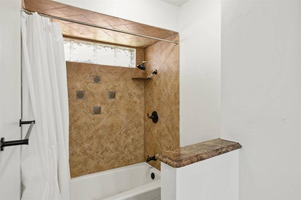 621 Harvest Glen Drive, Richardson, TX 75081 Shower and bathtub combination featuring diagonal stone-finish wall tile, decorative metal accent inlays, a built-in corner shelf, glass block window, and dark metal hardware