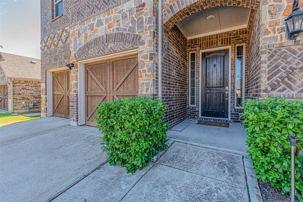 1742 Amalfi Drive, McLendon Chisholm, TX 75032 Exterior facade featuring a combination of brick and stone masonry, decorative brickwork accents, wood-finish carriage-style garage doors, a recessed arched entry, and a multi-paneled dark wood entry door with side windows