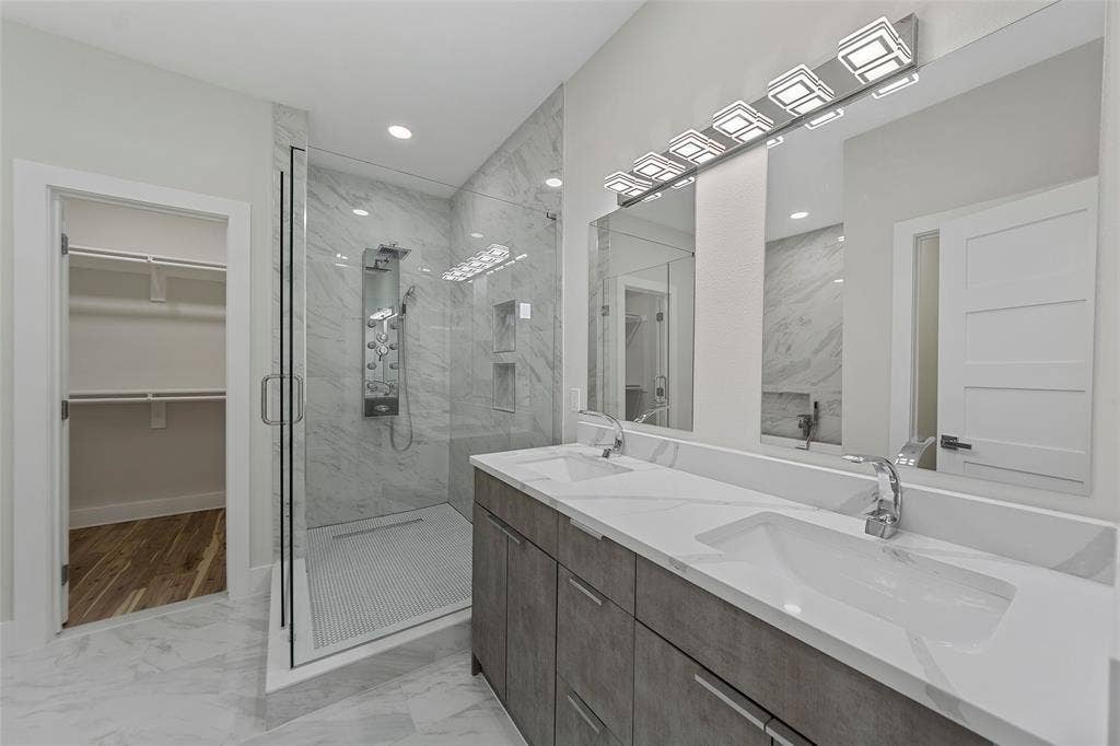 2503 Crossman Avenue, Dallas, TX 75212 Dual-sink vanity with white stone-surface countertop, walk-in shower with multi-head fixture, stone-style tile flooring, and integrated wall-to-wall mirrors
