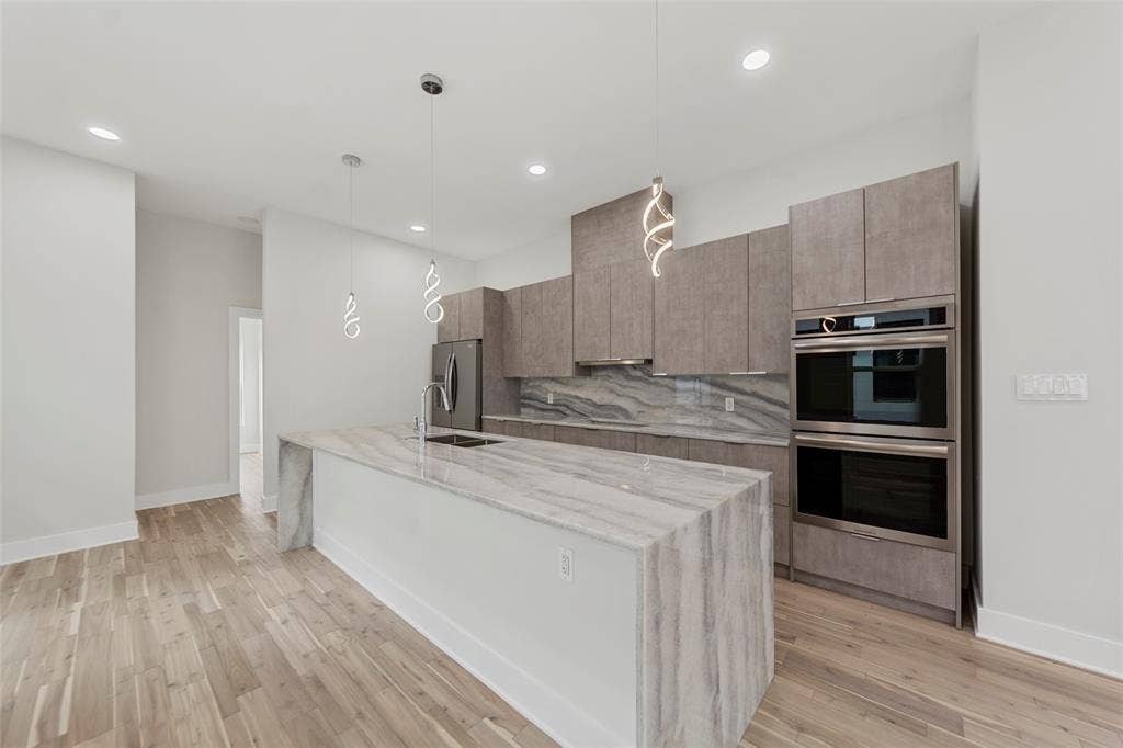 2503 Crossman Avenue, Dallas, TX 75212 Kitchen featuring a waterfall-edge island with stone surfacing, stainless steel double wall ovens, wood-finish cabinetry, patterned stone backsplash, and pendant lighting