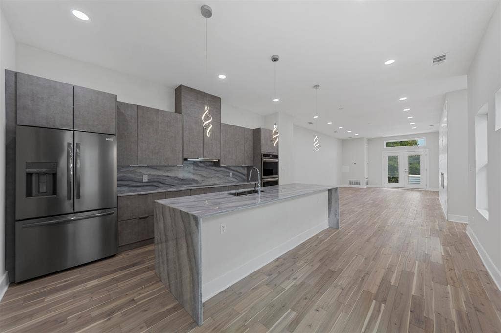 2503 Crossman Avenue, Dallas, TX 75212 Modern kitchen featuring a waterfall-edge stone island, flat-panel cabinetry, stainless steel appliances, pendant lighting, and wood-finish flooring