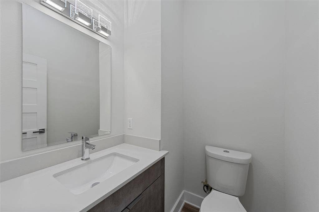 2503 Crossman Avenue, Dallas, TX 75212 Vanity with a solid-surface countertop and integrated sink, sleek dark-toned cabinetry, rectangular mirror, contemporary linear light fixture, and ceramic toilet
