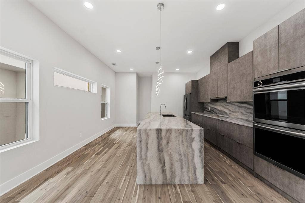 2503 Crossman Avenue, Dallas, TX 75212 Kitchen featuring an expansive waterfall-edge island, textured cabinetry, stone-patterned backsplash, wood-finish flooring, and integrated stainless steel double wall ovens