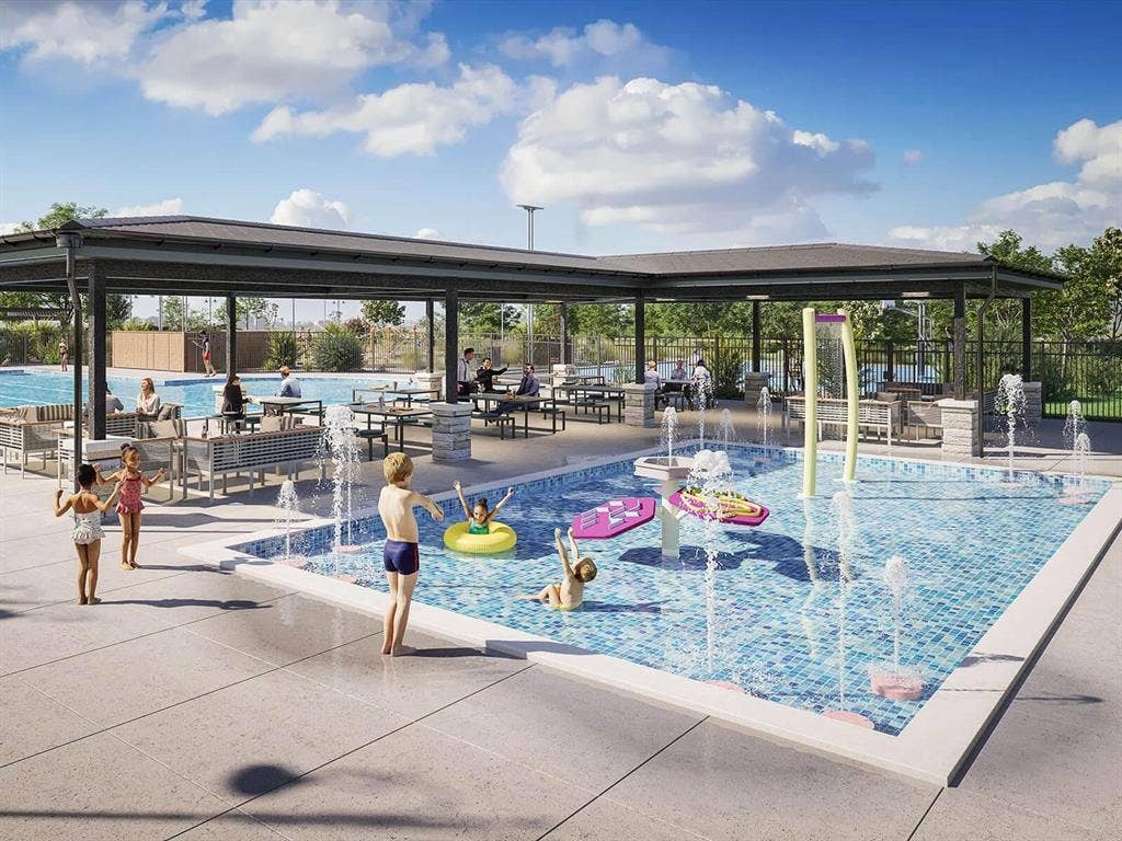 Outdoor aquatic area featuring a splash pad with water jets, adjacent swimming pool, and a large covered pavilion with stone-clad support pillars and concrete patio surfacing