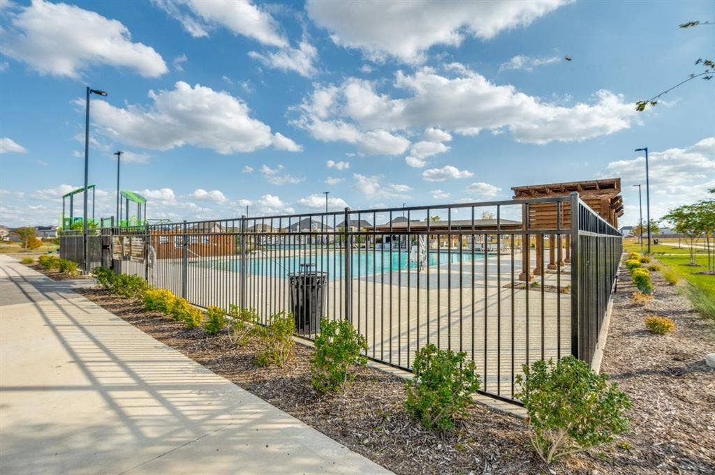Community swimming pool enclosed by metal fencing, featuring a wooden pergola and concrete patio surround