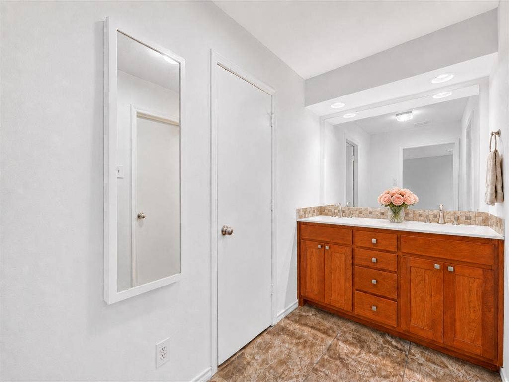 12627 Breckenridge Drive, #200, Dallas, TX 75230 Double vanity featuring stained wood cabinetry, a solid surface countertop, an integrated tiled backsplash, and a large-format mirror with recessed lighting