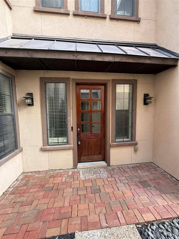 1025 Emerson Park Lane, Irving, TX 75063 Stucco exterior entry featuring a brick-paved patio, a metal-roofed awning, a wooden eight-lite front door, and wall-mounted exterior sconces