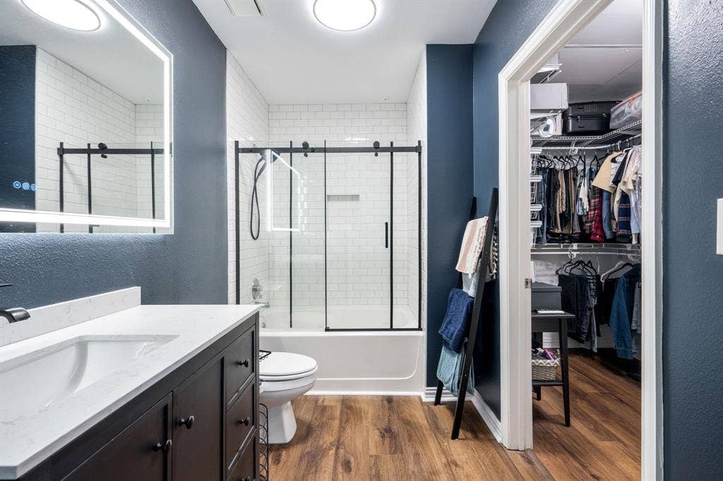 2305 Worthington Street, #103, Dallas, TX 75204 Bathroom featuring a vanity with a white countertop, an illuminated mirror, wood-finish flooring, a tub-shower combination with a black-framed sliding glass enclosure, and white subway tile wall surrounds
