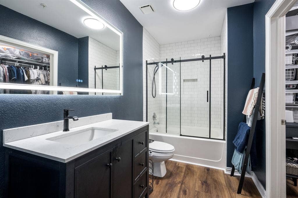 2305 Worthington Street, #103, Dallas, TX 75204 Bathroom featuring a dark wood vanity with a white stone-composite countertop, an integrated LED-backlit mirror, wood-finish flooring, and a bathtub with white subway tile surround and a sliding glass door with a black frame