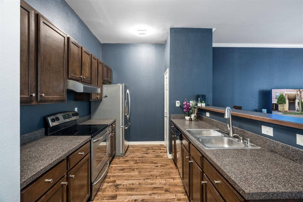 2305 Worthington Street, #103, Dallas, TX 75204 Kitchen featuring dark wood cabinetry, textured countertops, stainless steel appliances, a dual-basin stainless steel sink with a high-arch faucet, and wood-finish flooring