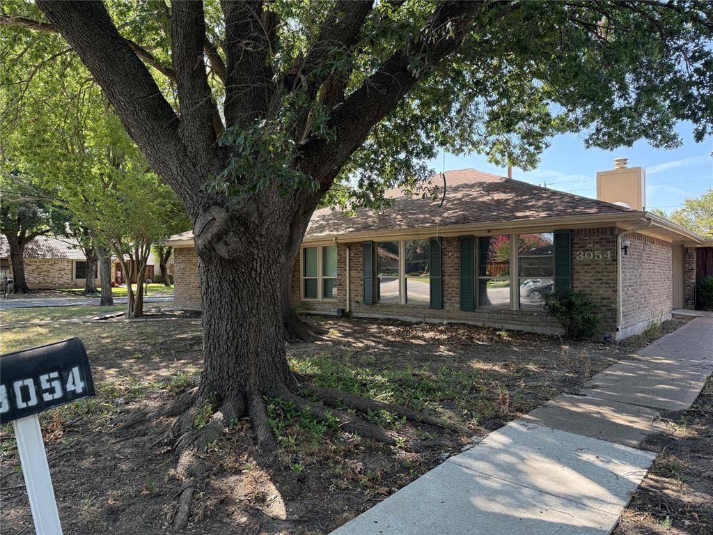 Single-story brick exterior featuring a hip roof, chimney, shutter-framed windows, and mature trees