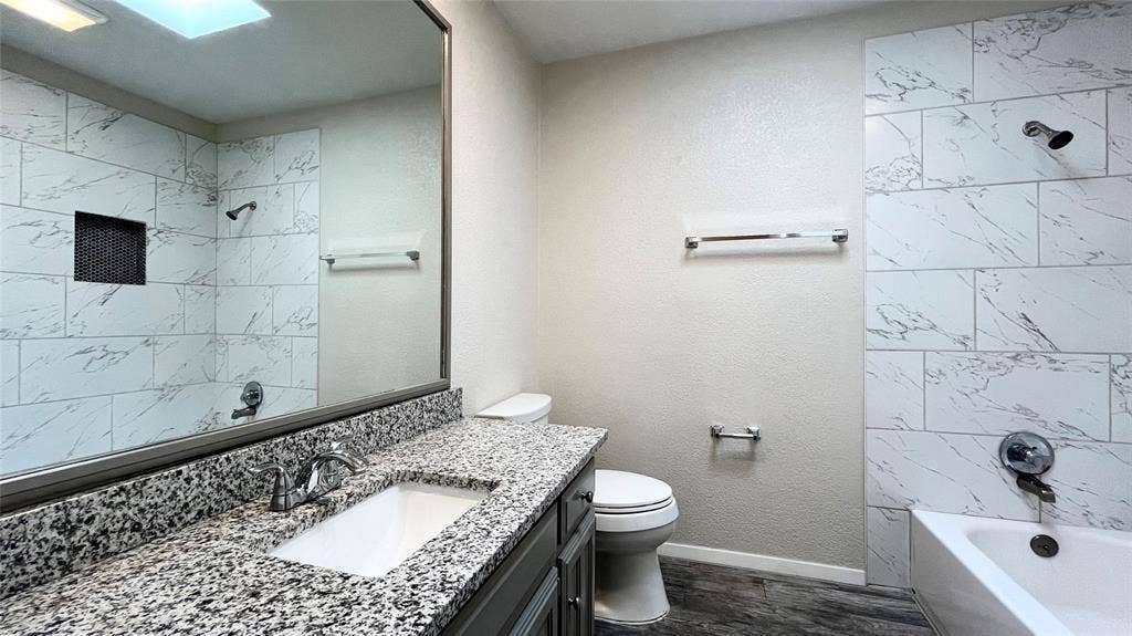 Full-width vanity with a stone-finish countertop, undermount sink, large wall-mounted mirror, bathtub featuring large-format tile surround, and built-in shower niche