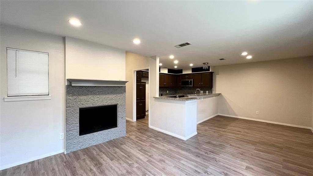 Fireplace with stacked-stone surround and mantel, wood-finish flooring, recessed lighting, and kitchen featuring dark cabinetry and a peninsula
