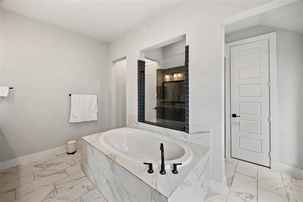 2820 Eccleston Street, Celina, TX 75009 Oval soaking tub set in a stone-finish deck with a matte black faucet, surrounded by large-format stone-finish floor tile, a tiled accent wall with integrated mirror, and a five-panel solid core door