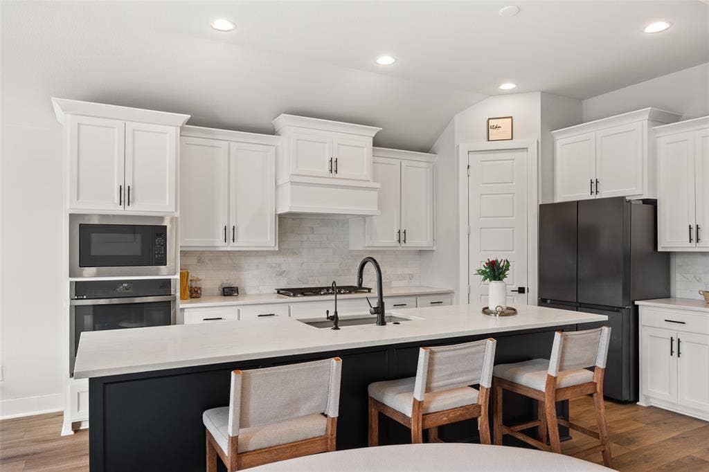 2820 Eccleston Street, Celina, TX 75009 Kitchen featuring white shaker-style cabinetry, a contrasting dark island with a quartz countertop, stone tile backsplash, built-in stainless steel appliances, and wood-finish flooring