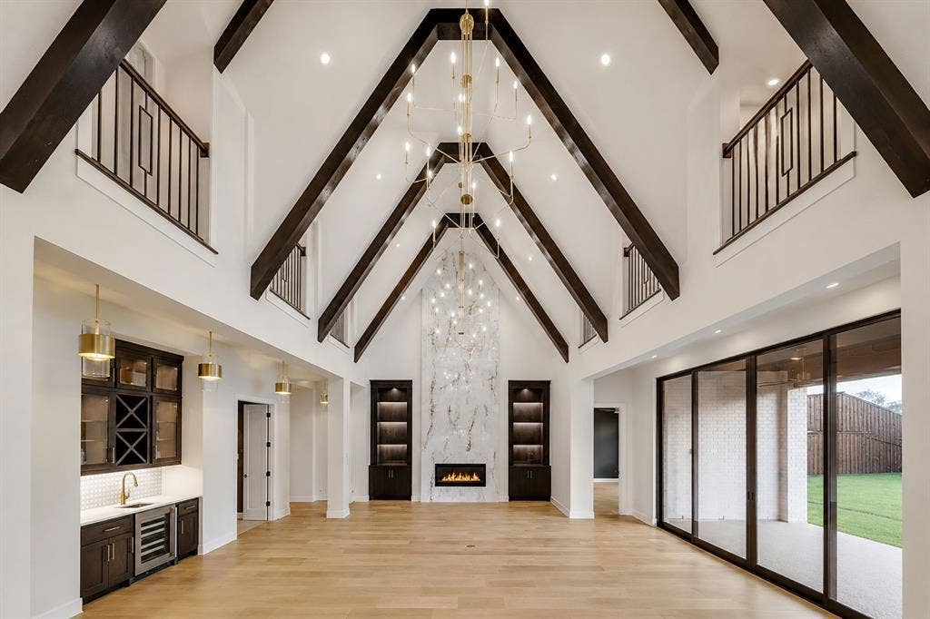 Vaulted ceiling with exposed decorative beams, floor-to-ceiling stone fireplace, built-in wet bar, multi-panel sliding glass doors, and mezzanine-level balconies