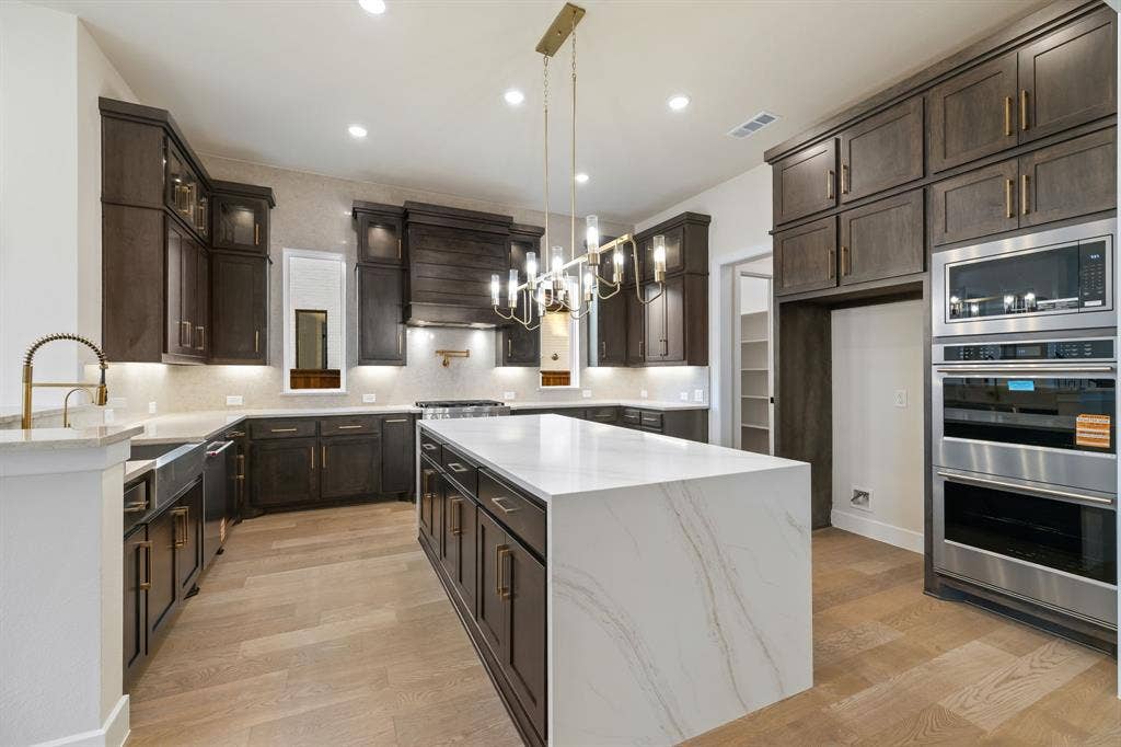 Kitchen featuring a central island with a solid surface countertop, dark-finish cabinetry, built-in stainless steel double wall ovens, wood-finish flooring, and a multi-light chandelier fixture
