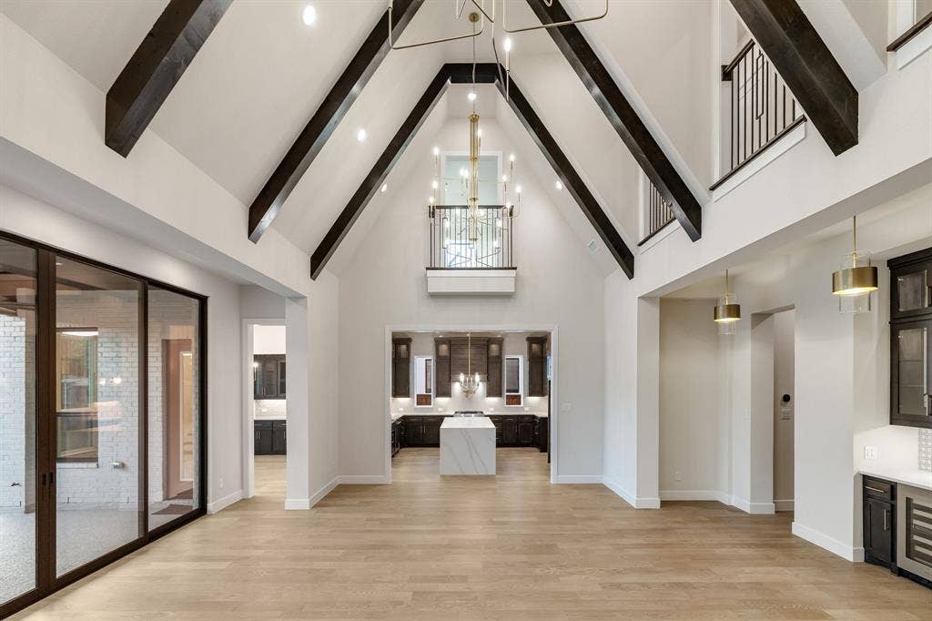 Vaulted ceilings with exposed beams, wood-finish flooring, an interior balcony with metal railing, floor-to-ceiling glass panel doors, and a kitchen island with a stone-finish countertop