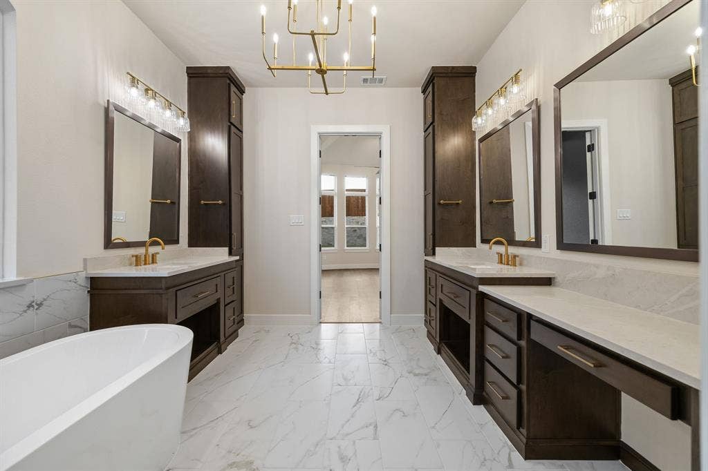Primary Bath Dual vanity area featuring dark wood cabinetry, light-toned stone countertops, brass-toned fixtures, a freestanding soaking tub, and large-format tile flooring