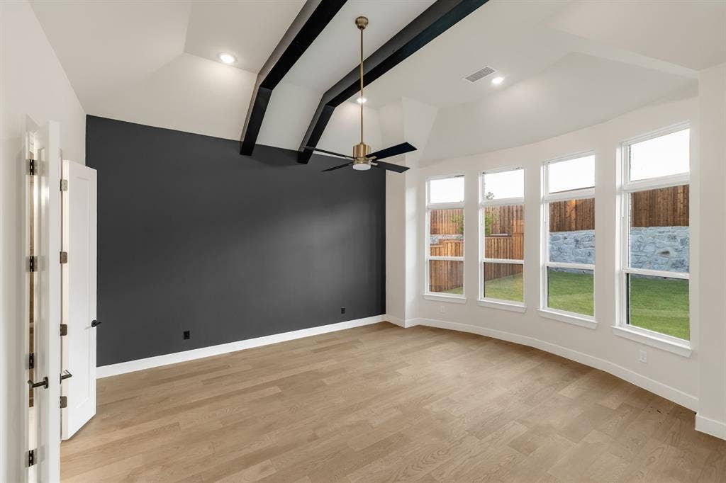 Vaulted ceiling featuring exposed dark-finish architectural beams, light wood-finish flooring, accent wall, and a series of windows
