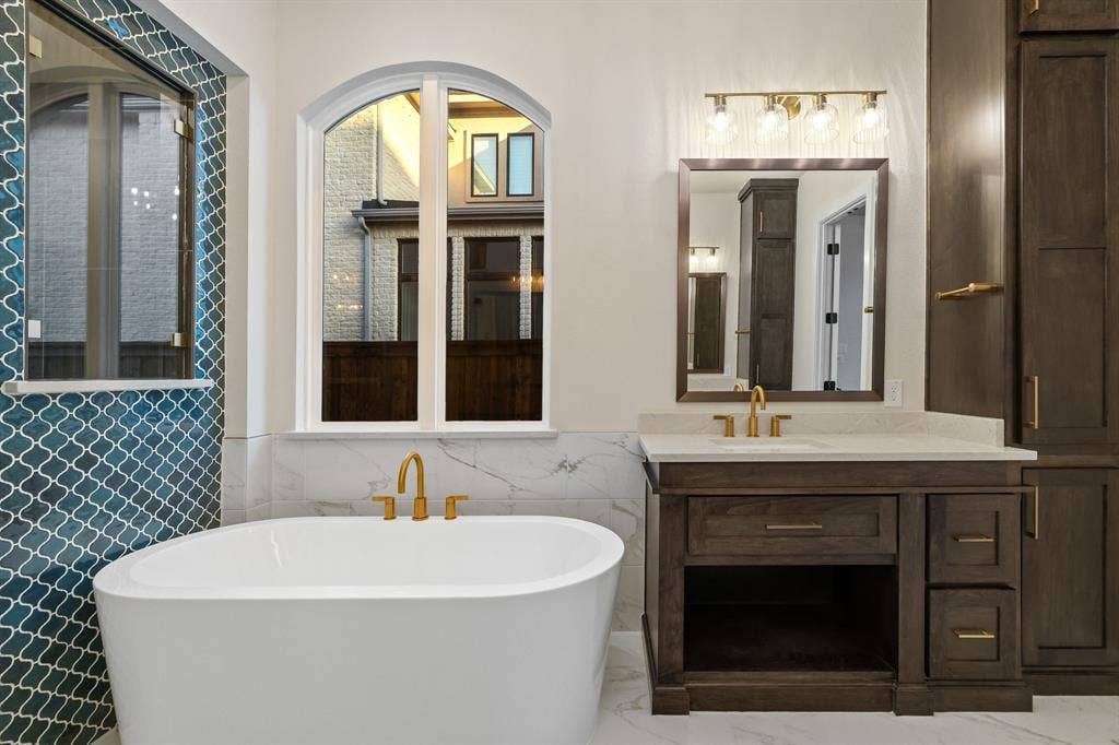 Primary Bath Freestanding soaking tub with a wall-mounted arched window, brass finish fixtures, decorative blue tile backsplash, and dark wood cabinetry with a stone countertop