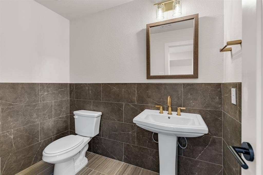 Upstairs Powder room featuring stone-finish wall tile, pedestal sink, gold-tone faucet hardware, framed mirror, and two-light wall sconce