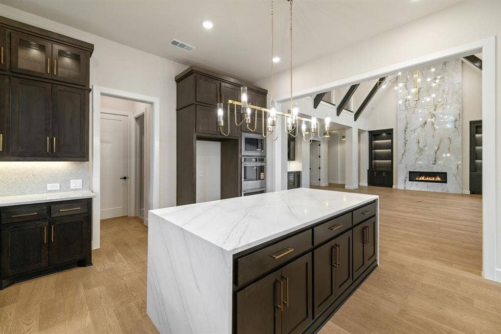Kitchen featuring dark-finish cabinetry, a waterfall-edge island with stone-finish countertops, brass-tone hardware, and a linear pendant light fixture, with wood-finish flooring throughout the space