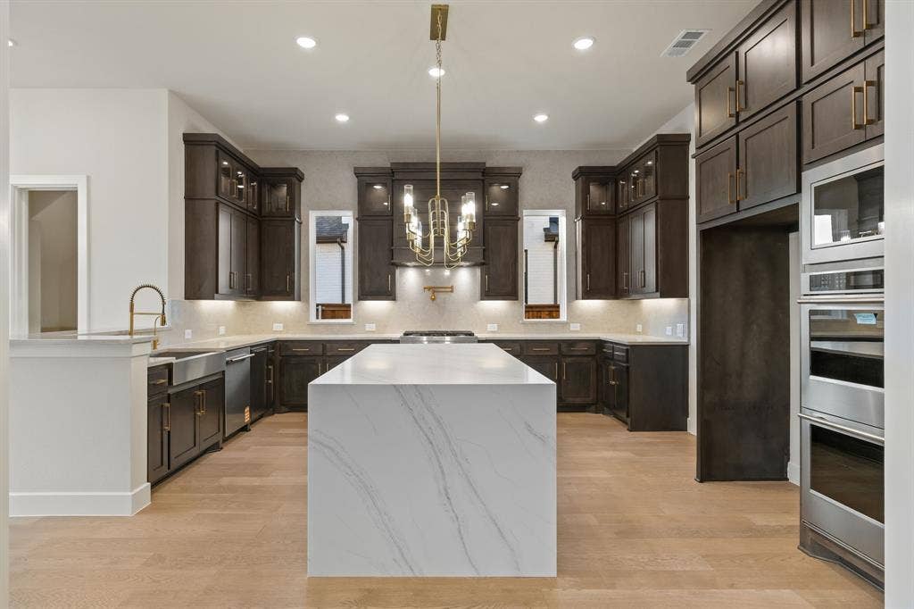 Kitchen featuring dark wood cabinetry, a large center island with a stone-finish waterfall edge, stainless steel appliances including a built-in double oven, wood-finish flooring, and a decorative metallic chandelier