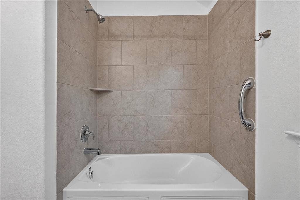 Bathtub alcove featuring neutral-toned tile surround, built-in shelving, chrome fixtures, and a wall-mounted support bar