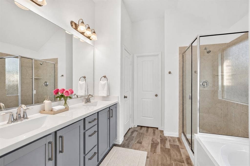 Dual-sink vanity with light-tone cabinetry, stone-surface countertop, full-width mirror, tile-surround corner shower, and wood-finish flooring