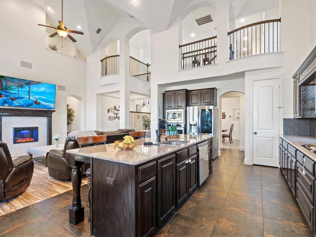 1506 Hennessey Drive, Allen, TX 75013 Kitchen with arched walkways, dark brown cabinets, open floor plan, light stone countertops, and high vaulted ceiling