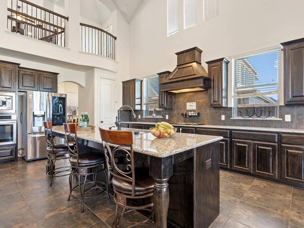 1506 Hennessey Drive, Allen, TX 75013 Kitchen featuring dark brown cabinetry, appliances with stainless steel finishes, light stone counters, a kitchen bar, and a kitchen island with sink