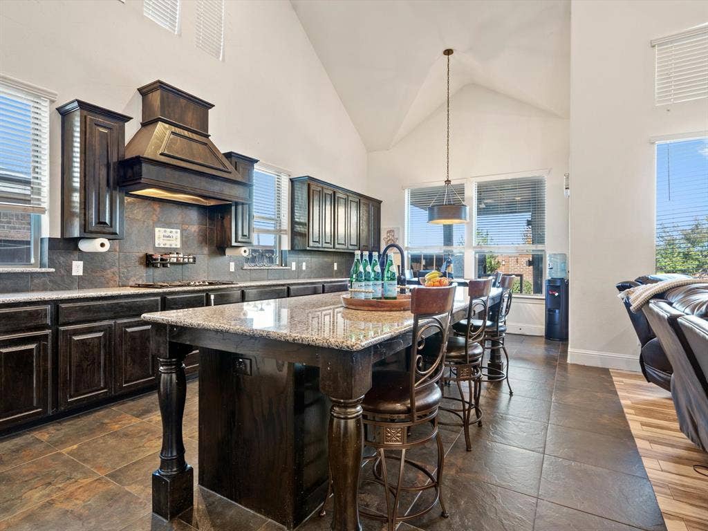 1506 Hennessey Drive, Allen, TX 75013 Kitchen featuring a kitchen breakfast bar, high vaulted ceiling, a kitchen island with sink, dark brown cabinetry, and light stone countertops