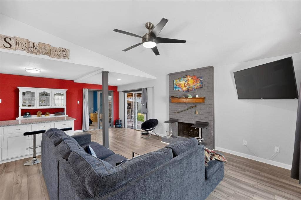 2228 Bunker Hill Drive, Carrollton, TX 75006 Living area featuring wood-finish flooring, vaulted ceilings, a grey painted brick fireplace with a wood mantel, a ceiling fan with integrated lighting, and a built-in wet bar with cabinetry